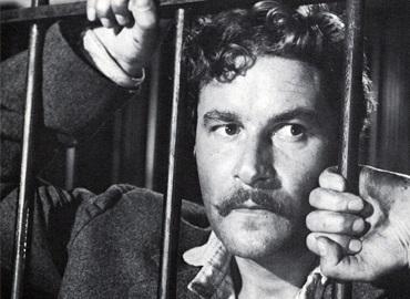 Black and white photo of man in prison looking forlorn, his hands on the bars