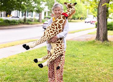 Anne Innis Dagg standing on a lawn wearing giraffe-print pants holding a large stuffed giraffe