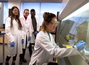 Sara Joffre in a lab coat and safety glasses using lab equipment as three of her classmates watch in the background