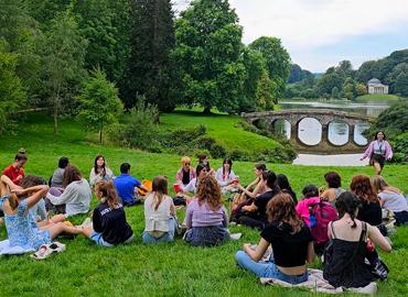A group of students sitting on green grass by a bridge and a river.