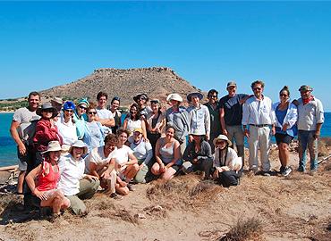 Professor Carl Knappett and a large group of students stand in front of blue water and a large, protruding land mass on the isle of Crete.