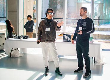 A man in VR glasses standing beside another man looking at him