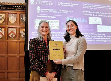 Professor Gillian Einstein presents award to PhD student Laura Gravelsins on a stage.