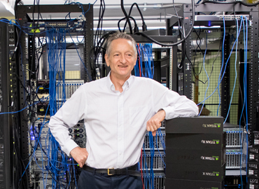 Geoffrey Hinton standing in front of a wall of computer servers.