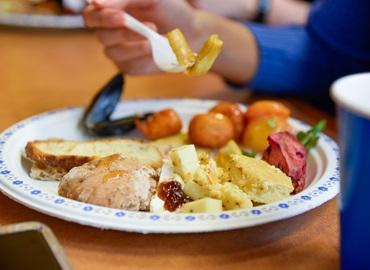 A person&amp;#039;s hand holding a fork above a paper plate loaded with different foods