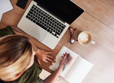 Overhead image of a woman near her laptop, writing in a notepad with a cup of coffee beside her.