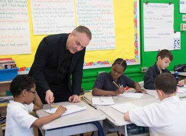 John Mighton in elementary classroom helping students at desks.