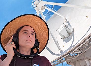 Kelly Lepo with a pair of headphones on listening beside a satellite dish.