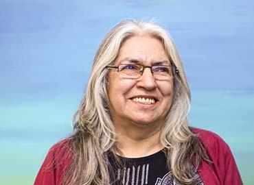 Headshot of Lee Maracle smiling wearing a red jacket and glasses