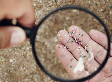 hands holding a maginifying glass to look at tiny pieces of plastics