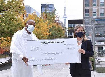 Bikila Award president Tessema Mulugeta presents A&amp;amp;S dean Melanie Woodin standing outside with masks on holding a large cheque.