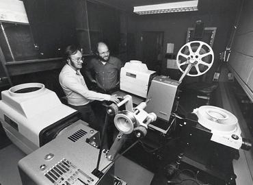 A black and white photo of David Clanfield (left) and Joe Medjuck behind a movie projector.