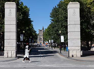 woman walking across a road with U of T gates to campus