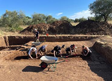 Five archaeologists digging in an open pit on a sunny day in Italy - a wheelbarrow is visible in the foreground