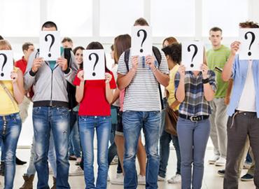 A group of students stand side-by-side. In front of their faces, they each hold a sign with a question mark on it.