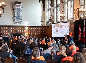 Brenda WastasecooT in an Orange Shirt at a podium in front of a seated audience in Hart House