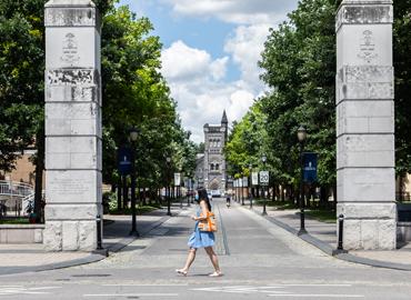 A person in a blue dress and blue medical mask walks on College st. with Kings College in the background on a sunny day.