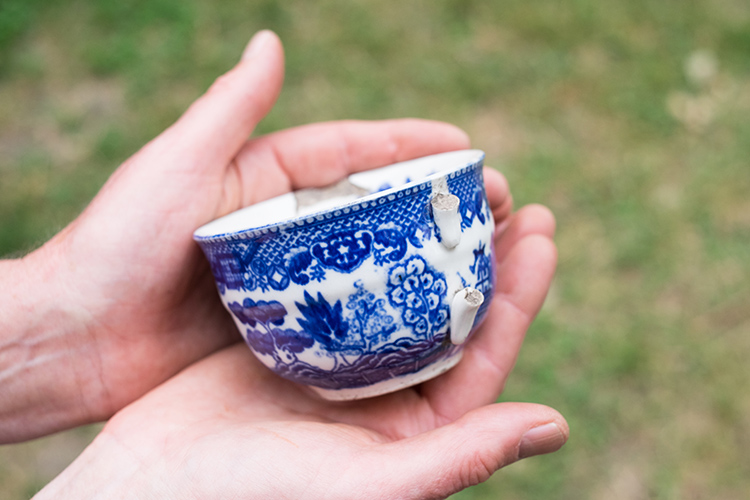 Two hands holding a teacup with a blue willow pattern and broken handle from circa in the mid-19th century.