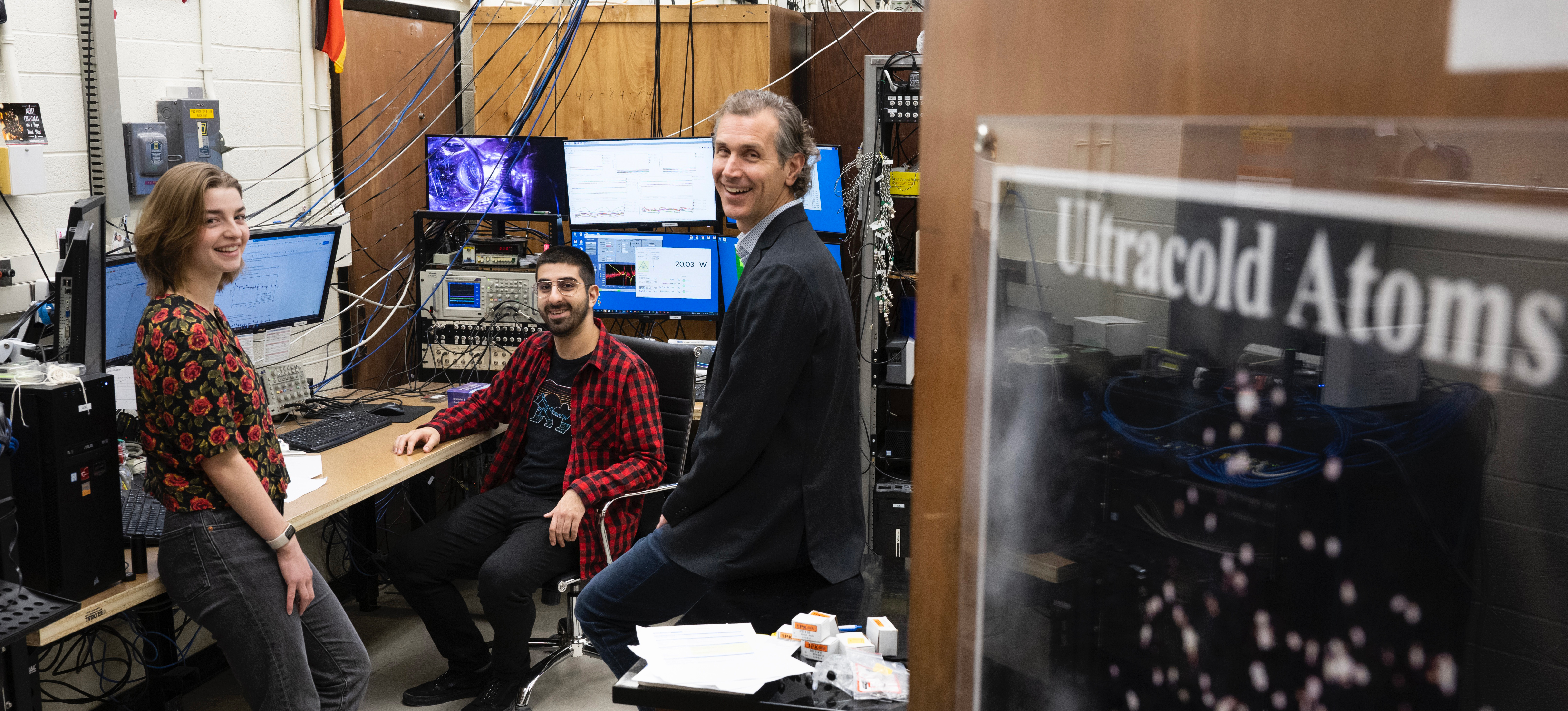 Robyn Learn, Frank Corapi and Joseph Thywissen standing in a lab with a sign on the door visible reading "ultracold atoms"