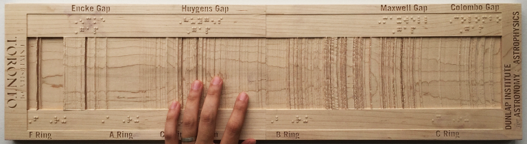 A hand touching a long wooden board carved with ridges representing Saturn’s main ring system
